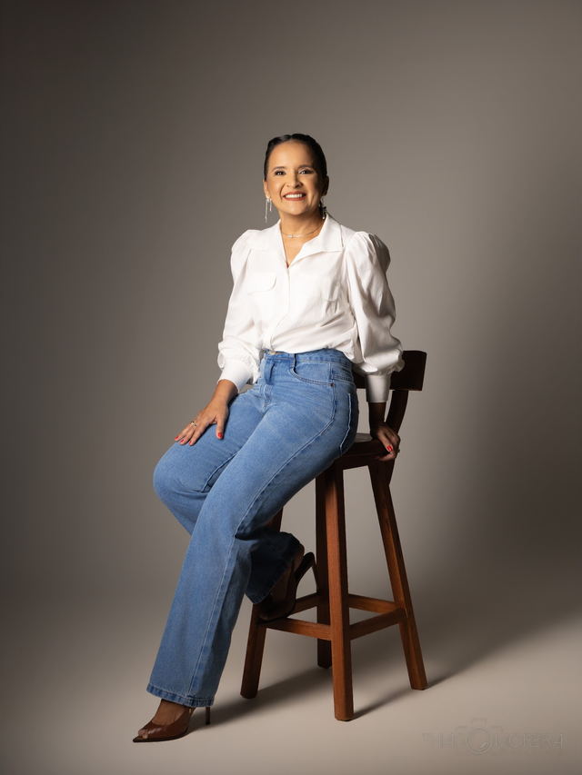 Mulher de camisa branca e calça jeans sentada em uma banqueta alta, sobre fundo liso marrom