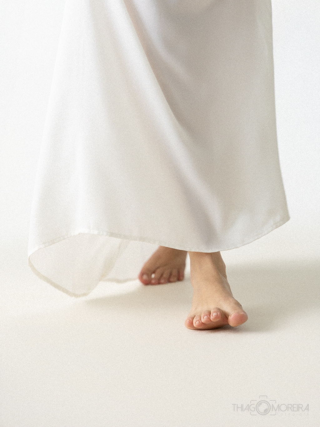 Detalhe dos pés descalços de uma gestante sob a barra de um vestido longo branco, em ensaio de estúdio branco sobre branco
