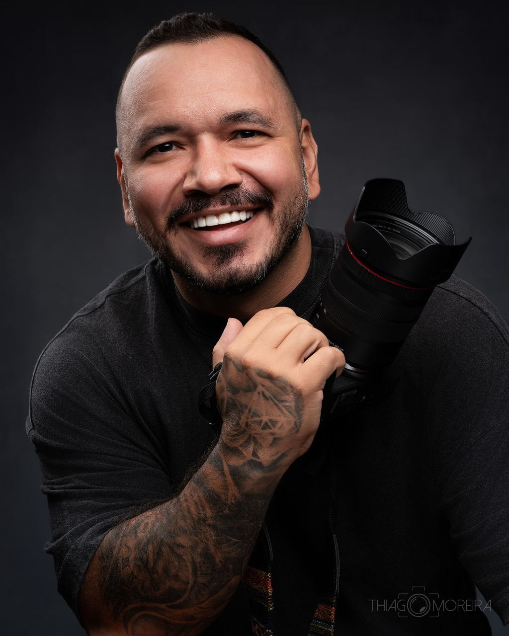 Thiago Moreira em retrato de estúdio sobre fundo escuro, sorrindo, com a câmera apoiada no ombro
